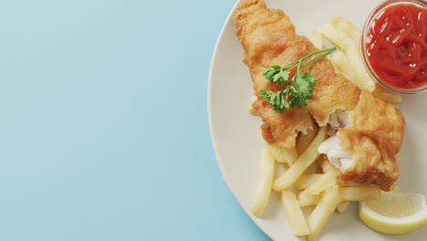 video close up of fish and chips, parsley and bowl of ketchup on plate, with copy space on blue