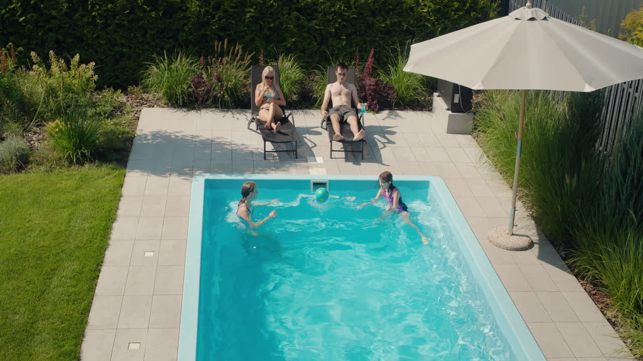 Familien slapper af ved deres pool i husets baghave. Voksne solbader på liggestole, to børn svømmer i poolen.
