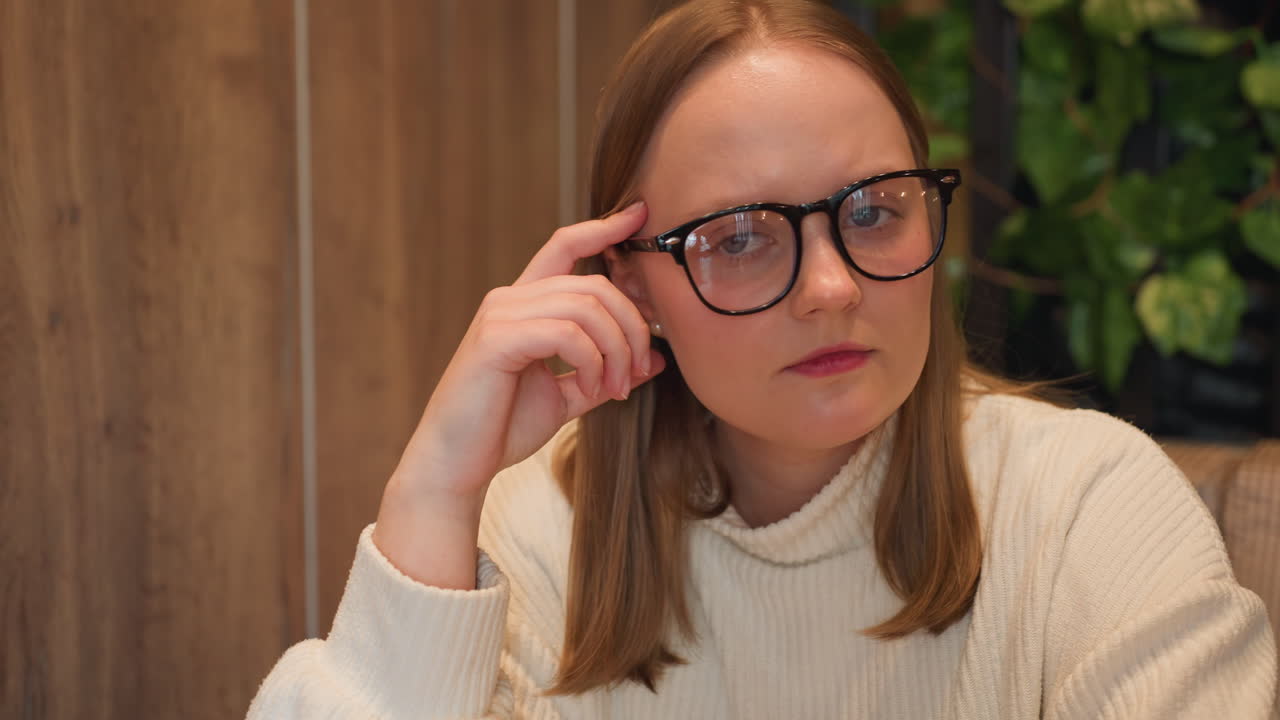 Close up of reflective young woman wearing glasses and white sweater, resting hand on temple while gazing thoughtfully, wooden wall background with leafy decor