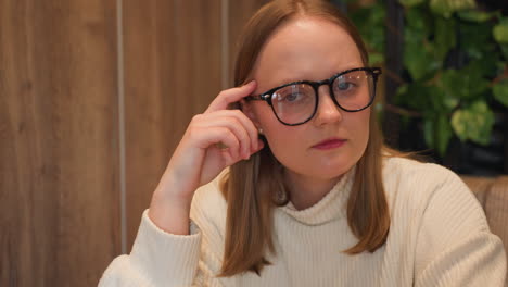 close up of reflective young woman wearing glasses and white sweater, resting hand on temple while gazing thoughtfully, wooden wall background with leafy decor