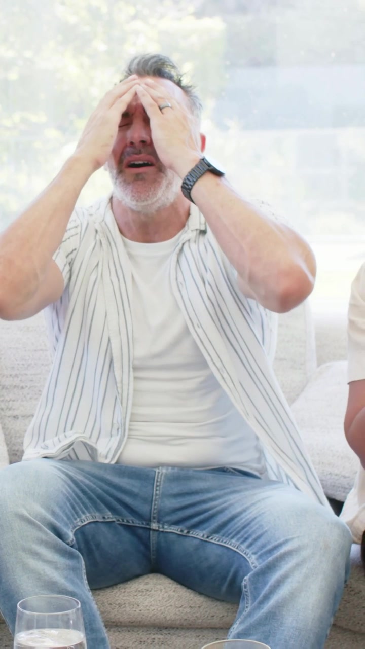 Vertical video: Speaking man in striped tee covering face, frustrated on couch, woman placing glass