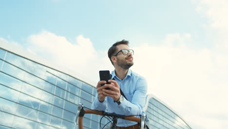 handsome man looking while holding in his hands a smartphone and sitting on the bicycle in the city