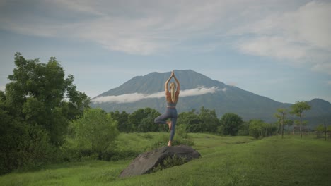 athletic blond woman in yoga tree pose balancing on rock, mount agung in background