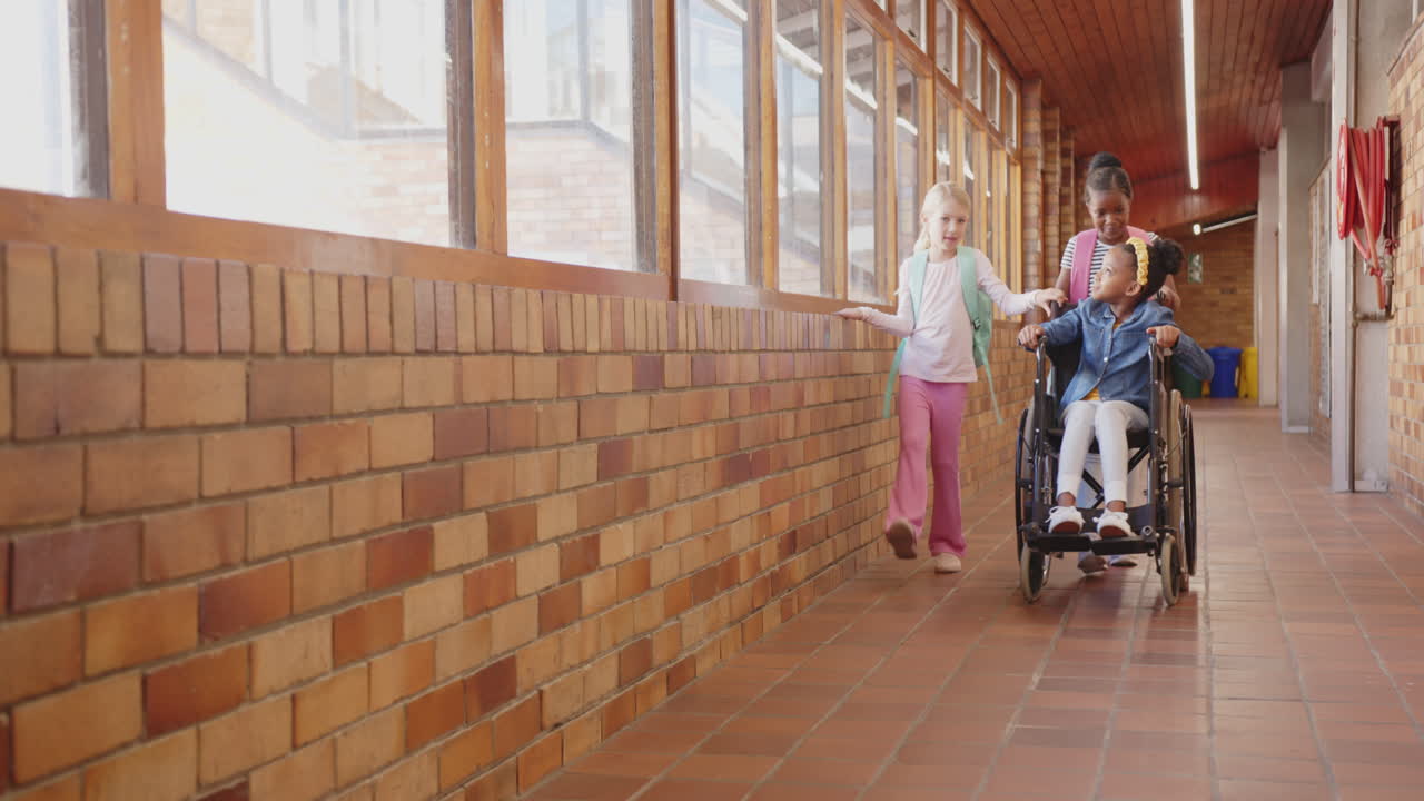 In school, two girls pushing friend in wheelchair down hallway, smiling and chatting