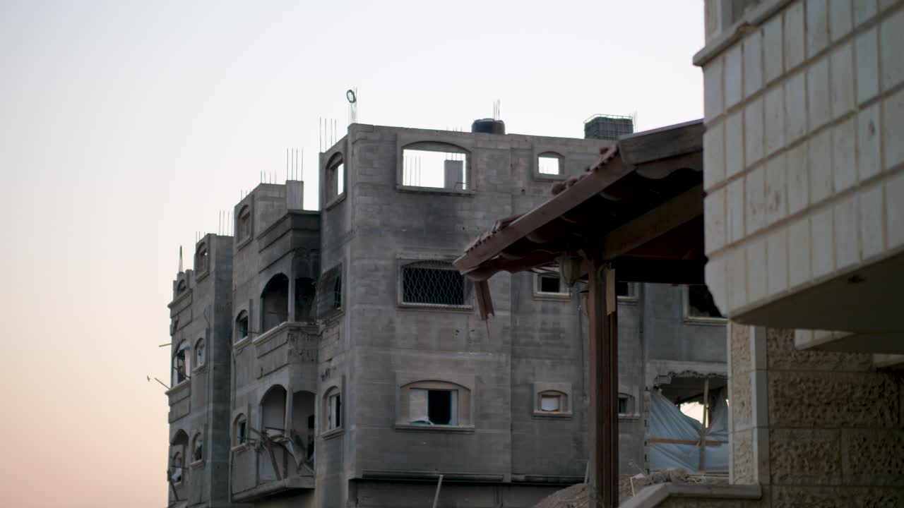 Inquadratura fissa interna attraverso un muro rotto che mostra edifici in rovina a Gaza.