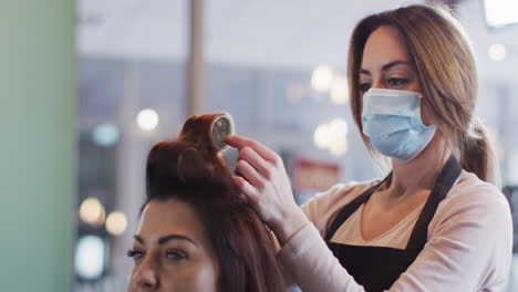 Female-hairdresser-wearing-face-mask-putting-rollers-on-hair-of-female-customer-at-hair-salon