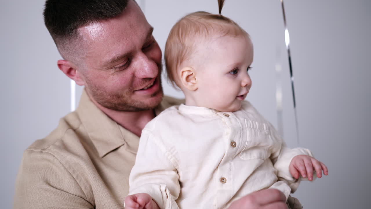 Bearded Caucasian man holding a lovely baby in hands. Active kid leans to catch the balloon. Close up.