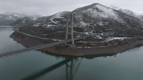 Bridge-long-road-crossing-wide-river-blue-winter-mountain-snowy-landscape-winter-skyline,-aerial-drone-zooming-into-infrastructure-at-natural-environment