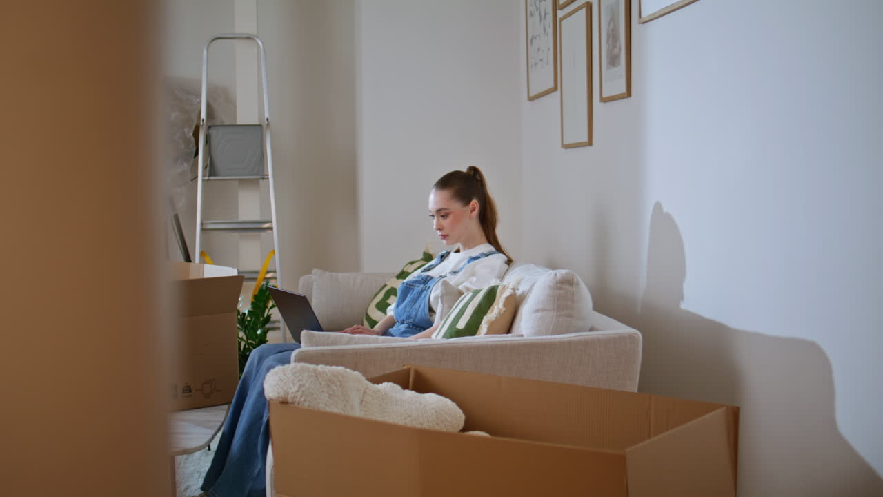 Moving girl shopping laptop online at comfortable couch new apartment slide view