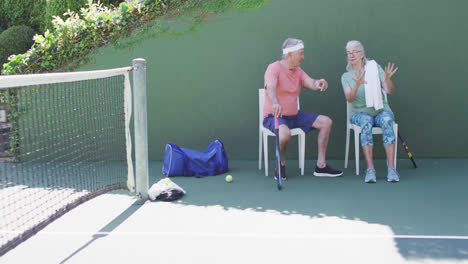 una feliz pareja caucásica senior tomando un descanso, sentada hablando en una cancha de tenis al aire libre, en cámara lenta