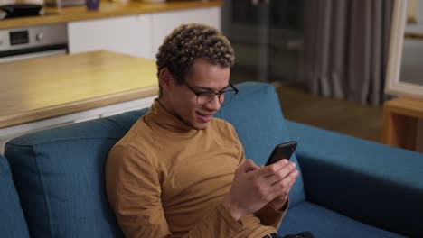 curly young man typing on his mobile phone on the sofa at home