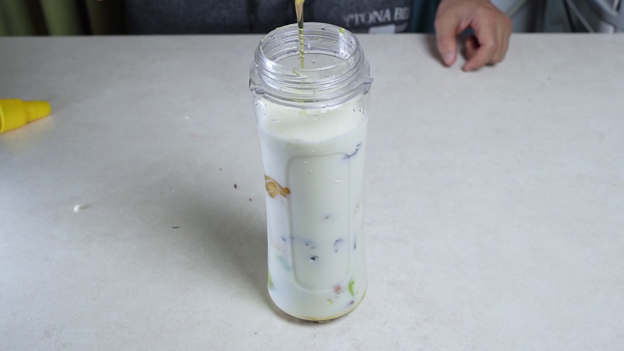 Lähikuva miehen käsistä puristamassa kultaista hunajaa muovipullosta kannettavaan tehosekoittimeen, joka on täytetty valkoisella maitosmoothiella ja hedelmillä. Ruoanvalmistus tapahtuu kirkkaalla keittiön työtasolla.