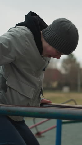close-up of a young boy smiling while attempting to climb an iron bar but unable to, he is dressed in a gray sweater and beanie, with a blurred background of people and buildings