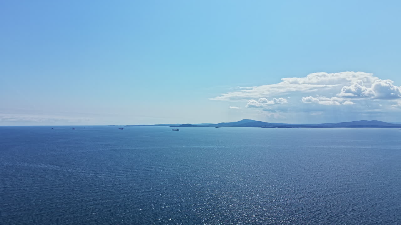 午後の澄んだ空の下、海岸線近くの青い海をヨットと貨物船がゆっくりと進む。