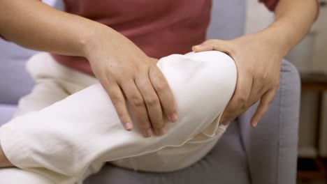 woman sitting on the sofa and massages her knee with her hands and examines her knee pain due to chronic walking aches and pains