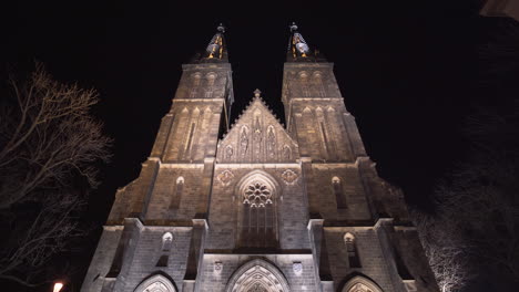 the illuminated entrance and front side of the gothic stone medieval church of saint peter and paul with its two spires at vyšehrad,prague,czechia,at night,on an empty paved square with lanterns,tilt