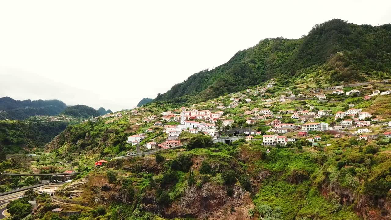A stunning view of Faial, Madeira, featuring a vibrant village nestled among lush green hills, dramatic cliffs, and winding roads, capturing the island's unique blend of nature and culture