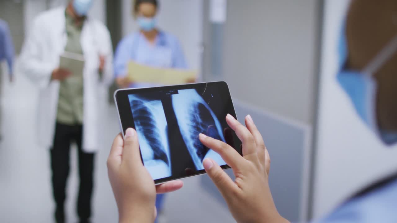 médico de raza mixta con máscara facial mirando rayos x en la tableta caminando en el pasillo del hospital