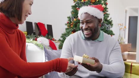 Video-of-happy-diverse-couple-in-santa-hats-exchanging-christmas-gift-and-embracing-at-home