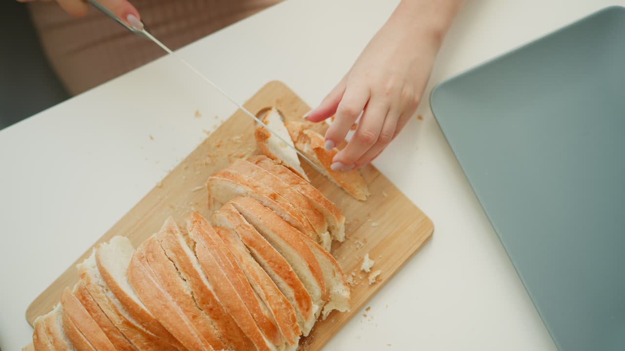 Close up hand view of young lady holding firm loaf of bread and slicing last piece with knife on neat white table under bright morning light, highlighting crisp crust and soft interior texture