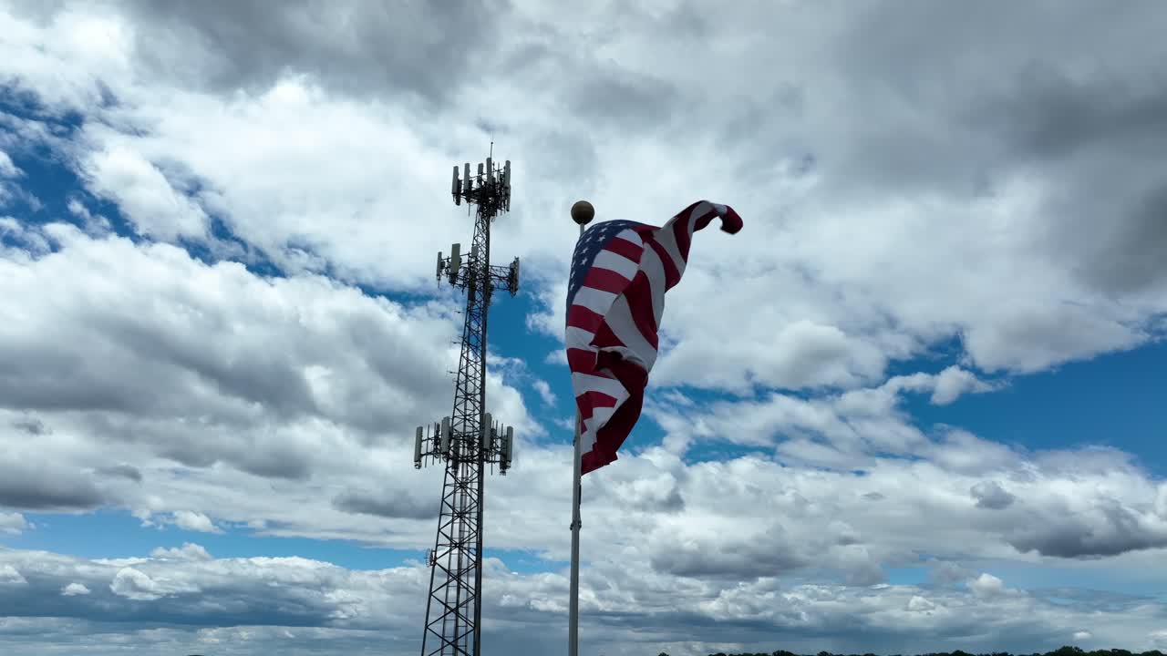 Waving american flag on flagpole in front of Transmission tower and dense clouds at sky. Bottom up shot. Proud and pride american country state.