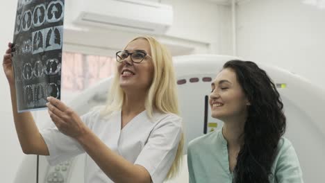 woman doctor radiologist explains good results of ct scanning for young female patient, showing the snapshot with images, observing and analyzing ct scan in modern clinic beside modern ct scanner