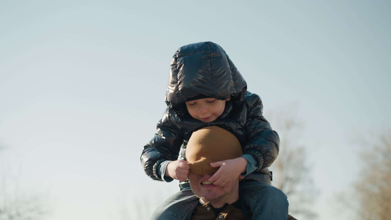 A close-up of a father carrying his young son on his shoulders while walking outdoors, the son playfully adjusts the father's beanie, which covers the father's face