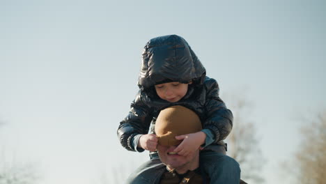 a close-up of a father carrying his young son on his shoulders while walking outdoors, the son playfully adjusts the father's beanie, which covers the father's face