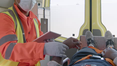 male paramedic with face mask using tablet computer and taking the pulse of an injured american patient who lying on the ambulance stretcher