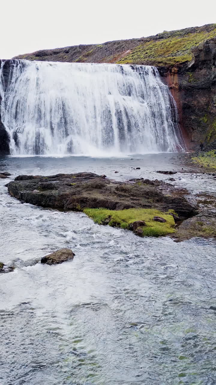 Aerial drone view of a large waterfall at the end of a river channel surrounded by moss covered cliffs and rocky slopes in Iceland's remote landscape. Vertical