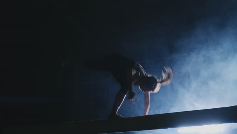 Side-view-of-a-female-gymnast-doing-split-handstand-on-balance-beam-against-black-background.