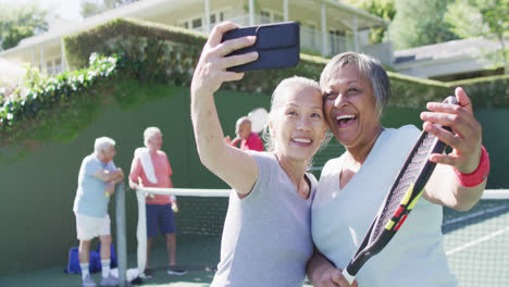 Dos-Felices-Y-Diversas-Mujeres-Mayores-Tomándose-Selfie-En-Una-Soleada-Cancha-De-Tenis-Al-Aire-Libre-Con-Amigos,-En-Cámara-Lenta