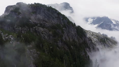 Aerial-dolly-through-wispy-clouds-as-forest-grows-up-along-rocky-cliff-in-PNW,-white-snow-and-green-vegetation-contrast-on-landscape