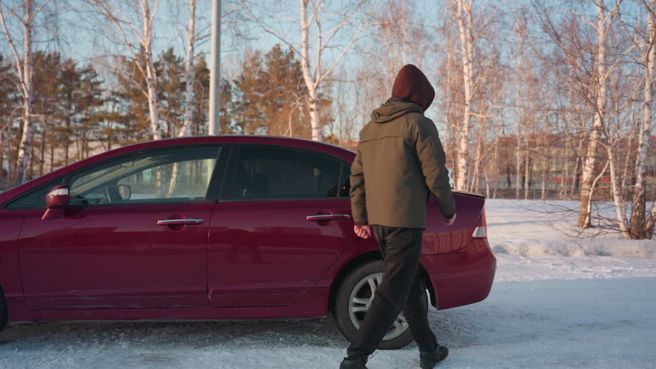 Man in hooded jacket opens red car driver-side door to access bonnet release in snowy outdoor parking lot with bare trees and daylight reflections on vehicle surface