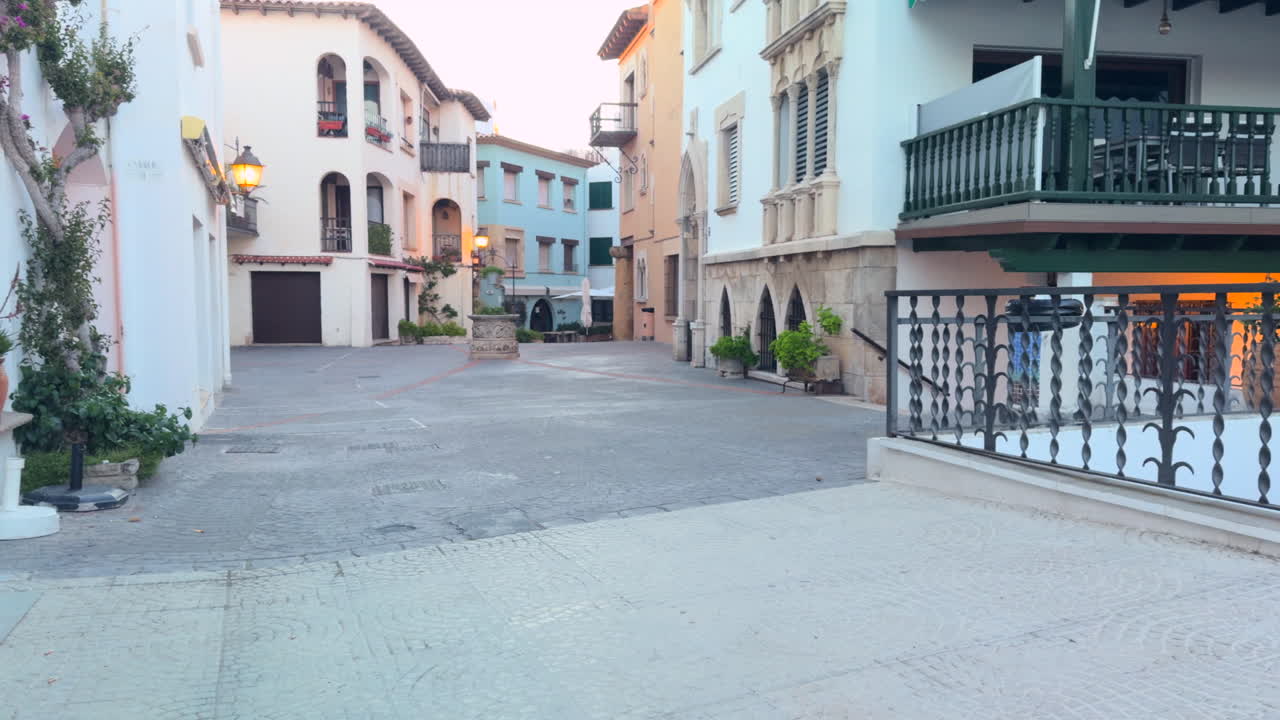 Scenic view of a quiet Mediterranean plaza surrounded by vibrant facades, wrought iron balconies, arched windows and lush plants in soft evening light. No people, calm atmosphere