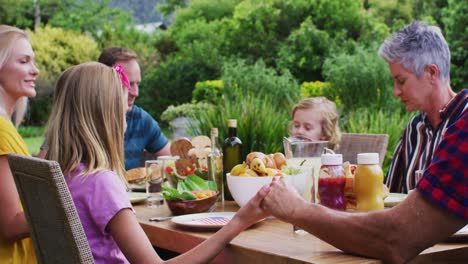 Smiling-caucasian-family-holding-hands-saying-grace-before-celebration-meal-together-in-garden
