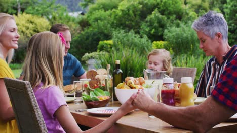 smiling caucasian family holding hands saying grace before celebration meal together in garden