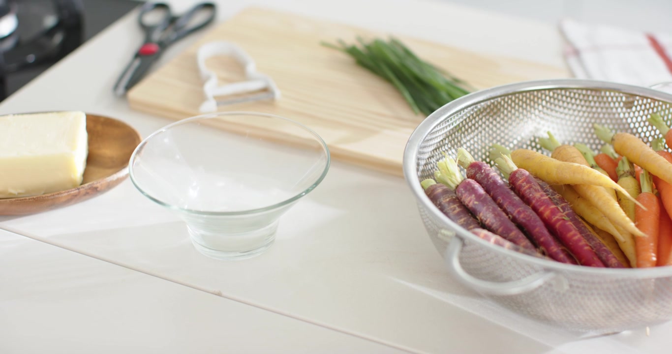 Camera is panning, revealing colander of multicolored carrots, glass bowl, butter, chives for prep