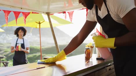 Hombre-Afroamericano-Con-Delantal-Y-Guantes-Limpiando-El-Camión-De-Comida-Con-Spray-Desinfectante