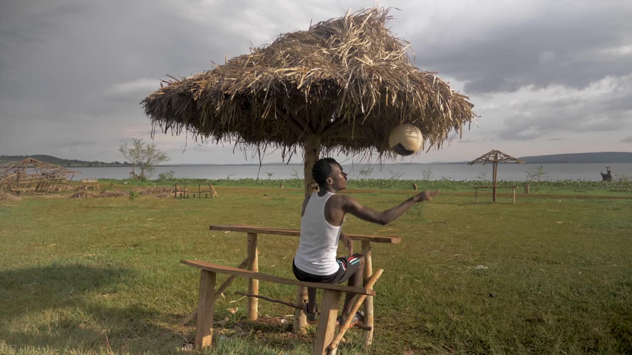 toma en cámara lenta detrás de un joven africano mientras se sienta bajo un refugio de cabaña en la playa mientras juega con una pelota de fútbol