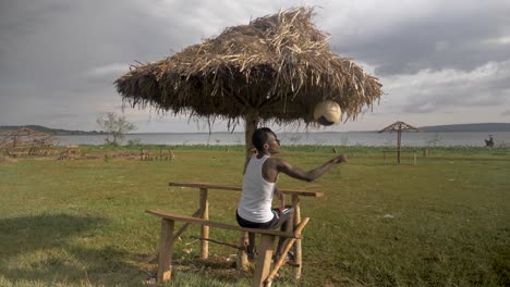 Toma-En-Cámara-Lenta-Detrás-De-Un-Joven-Africano-Mientras-Se-Sienta-Bajo-Un-Refugio-De-Cabaña-En-La-Playa-Mientras-Juega-Con-Una-Pelota-De-Fútbol