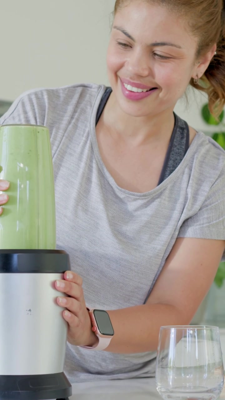 Video verticale: A casa, lei mette la tazza sulla base, la blocca e frulla un frullato verde, indossando abbigliamento sportivo.