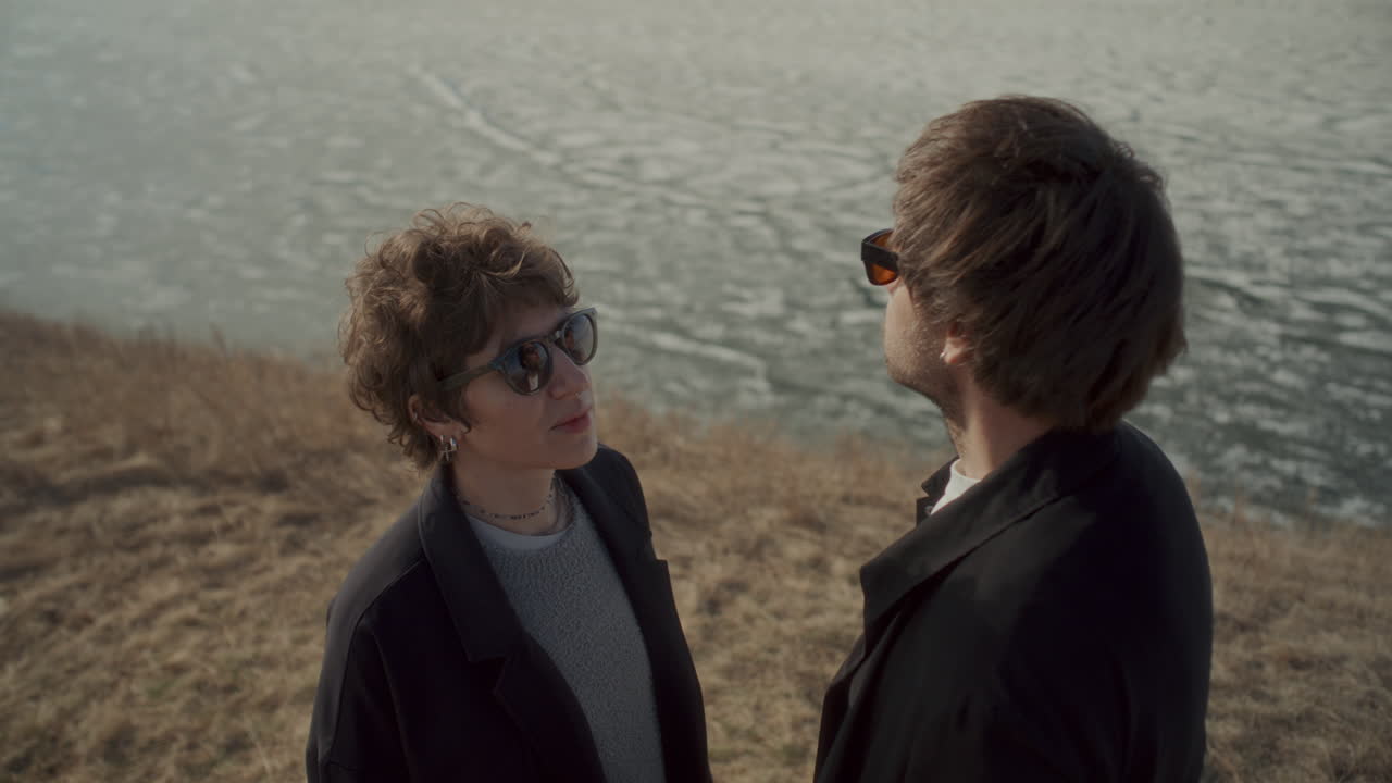 Couple in Sunglasses Standing on Lakeside Slope, Looking at Each Other