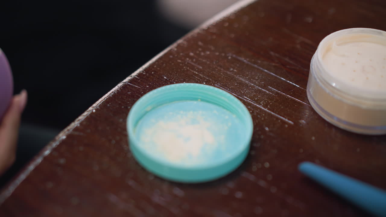 젊은 여성이 손으로 보라색 스폰지를 칠색 파우더 용기에 는 근접한 모습, 부드러운 초점으로 테이블에 배열 된 화장품 제품, 아름다움 일상을 보여줍니다.