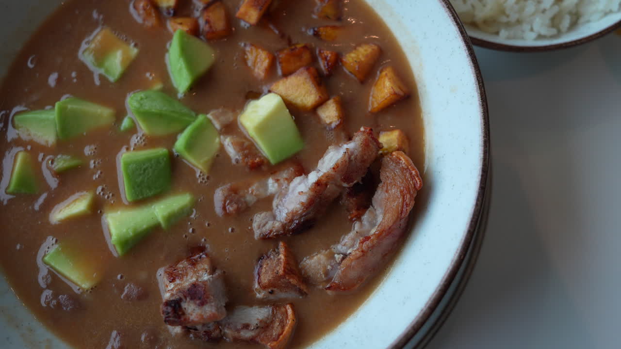 Close-up de uma cazuela de frijoles tradicional colombiana com pedaços de abacate, chicharrón crocante e acompanhamento de arroz. Capturada em iluminação suave para uma sensação reconfortante e acolhedora