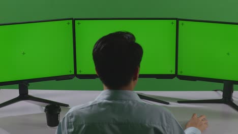 녹색 스크린 스튜디오에서 모의 여러 컴퓨터 모니터와 함께 작업하는 동안 스트레칭하는 피곤한 아시아 남자의 뒷면