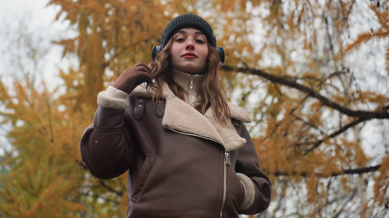 Thoughtful girl wearing black knit cap and brown shearling jacket with one hand in pocket, other hand rolling her hair outdoors during autumn day