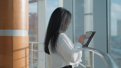 side view of investor in white shirt standing by iron railing holding tablet, overlooking urban skyline through large glass windows with bright daylight and reflections on transparent surfaces