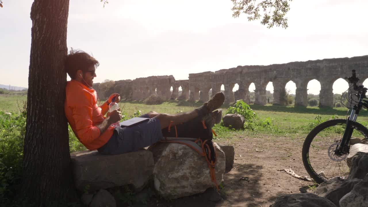 고대 로마 수로 앞에서 노트북과 함께 음악을 듣고 나무 의 바위 위에 앉아 있는 자전거 타는 사람