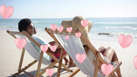 animación de corazones moviéndose sobre una pareja diversa enamorada en sillas de sol en la playa en verano